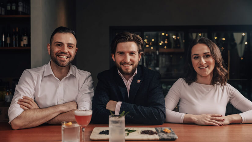 The three founders of Brunswick Aces sit at a bar smiling looking to camera