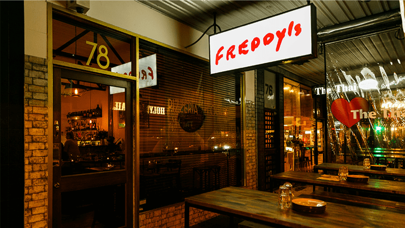 The shopfront of Freddy's pizzeria. A white and red neon sign saying "Freddy's" glows above outside tables set for dinner.