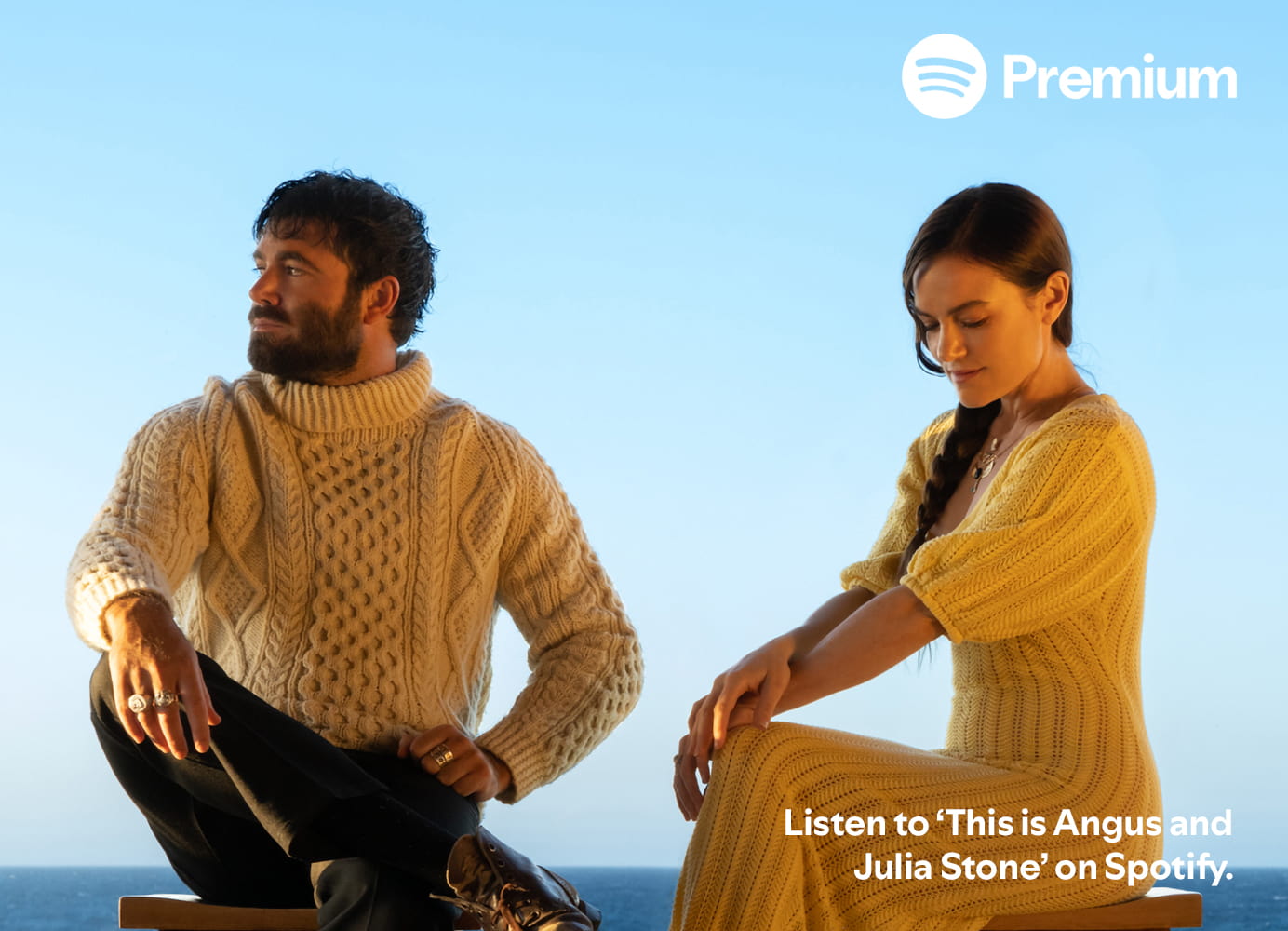 Two individuals are seated on chairs facing the ocean, with their backs to the viewer. One individual is wearing a white cable knit sweater and dark pants, while the other is in a yellow knit dress. The Spotify Premium logo is visible in the upper left corner against a clear blue sky. Text in the bottom right of the image is "Listen to 'This is Angus and Julia Stone' on Spotify."