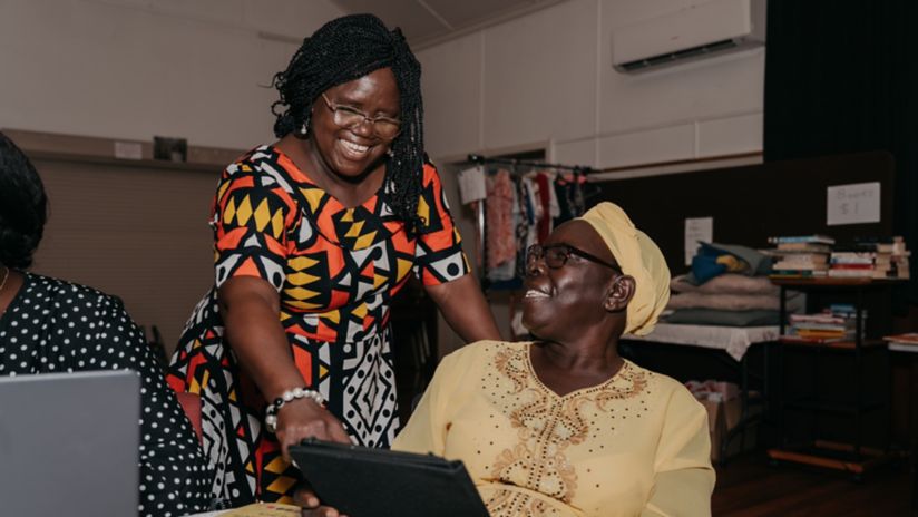 Two migrant women smile as they look at a tablet together, one seated and the other standing and pointing at the screen.