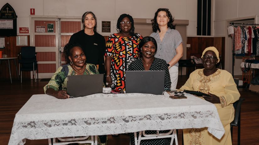 Six women in a group shot. Three are seated with laptops and a tablet, while the other three stand behind them, all smiling at the camera.