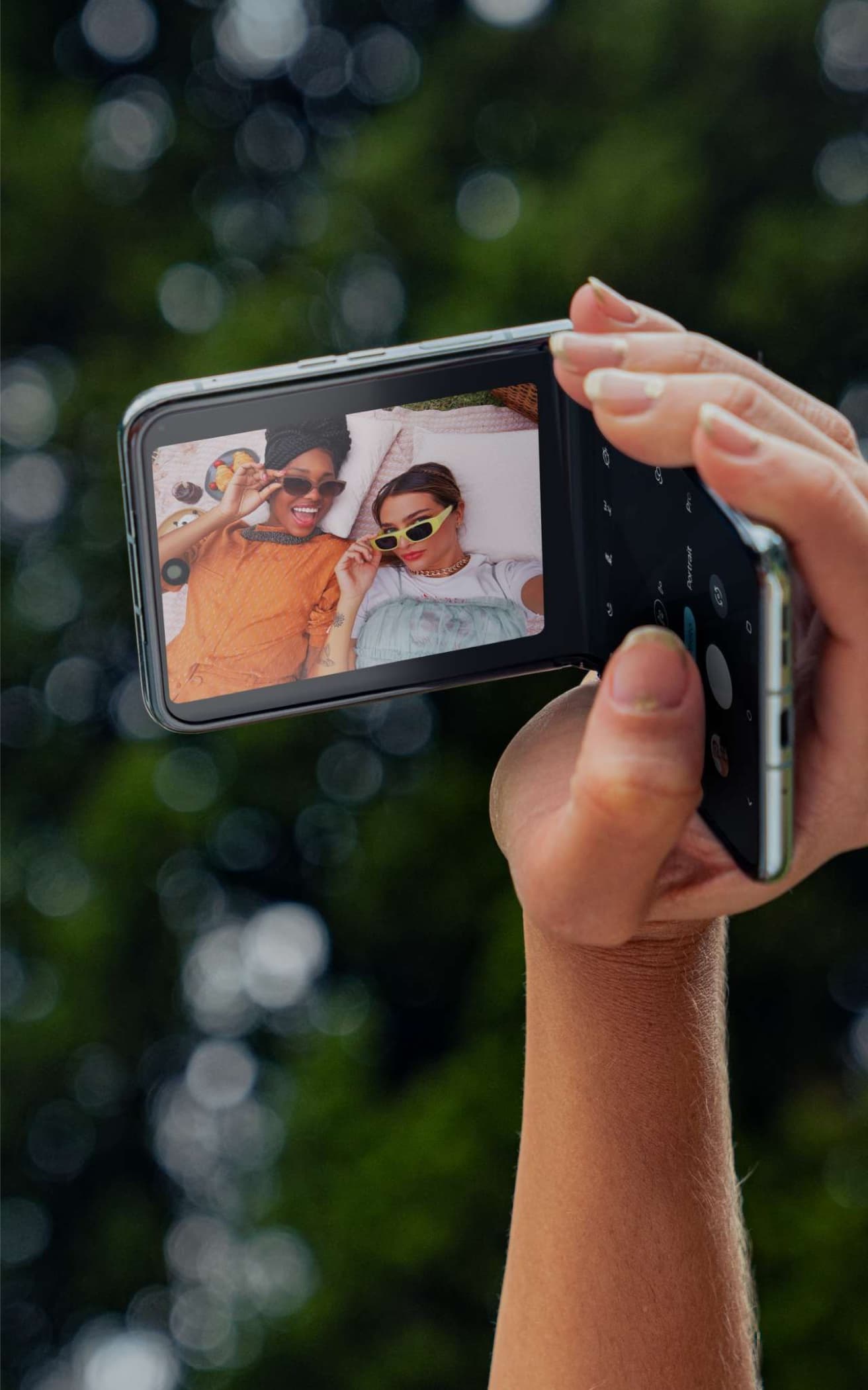 A hand holds a motorola razr 40 ultra flipped open to 90 degrees. The screen shows two happy people posing for a selfie.