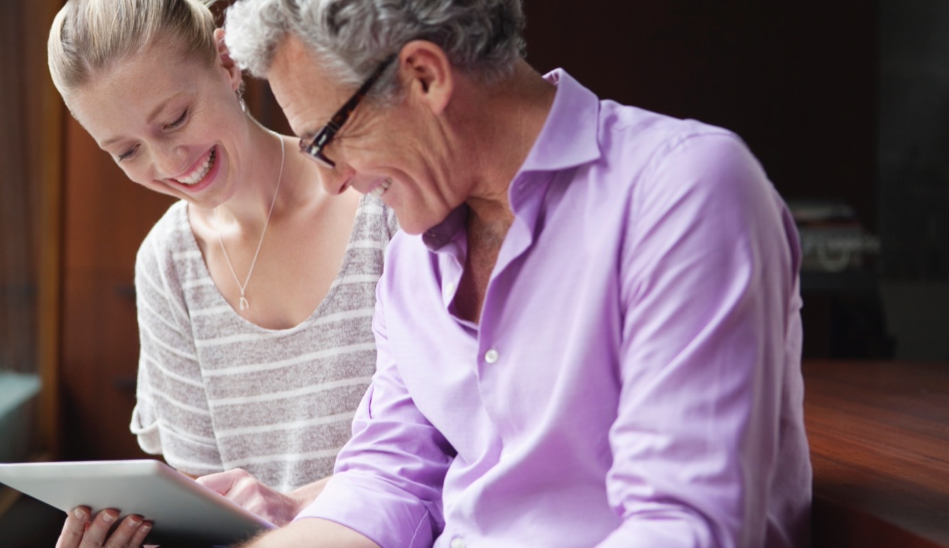 Two adults smile as they look over a tablet that one of the adults is holding. Only the rear side of the tablet is visible. 