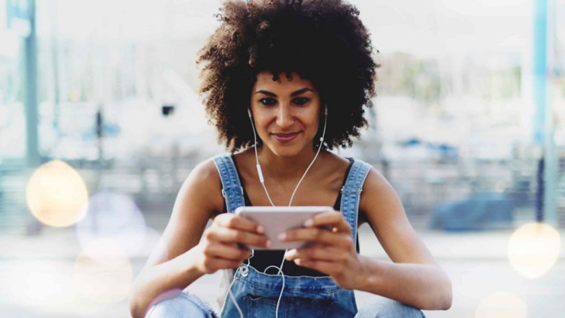 A young woman smiles as she looks down at her smartphone while wearing earbuds.