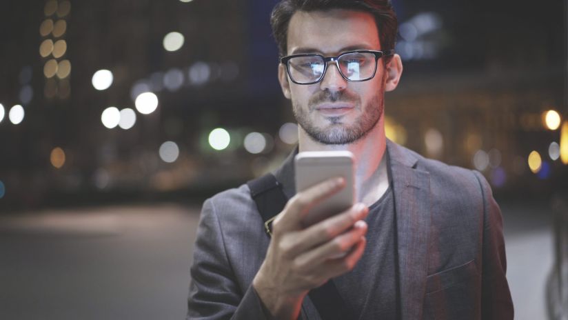 A man walking through city streets at night looks at his smartphone as he walks, and it illuminates his face with light.